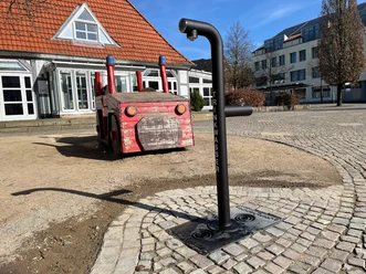 Schwarzer Trinkwasserbrunnen auf einem gepflasterten Platz neben einem Holz-Feuerwehrauto.