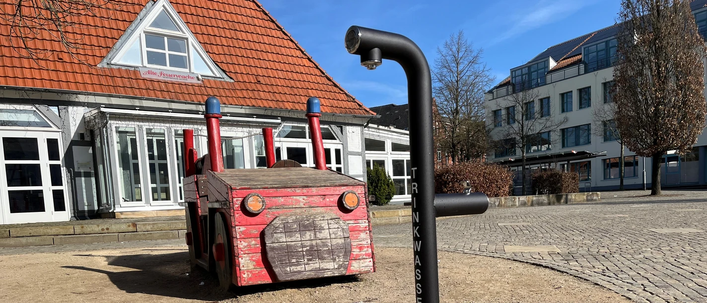 Schwarzer Trinkwasserbrunnen auf einem gepflasterten Platz neben einem Holz-Feuerwehrauto.