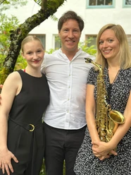 Summer Sounds.png Drei lachende Musiker, ein Mann und zwei Frauen, stehen draußen nebeneinander. Eine hält ein Saxophon.
