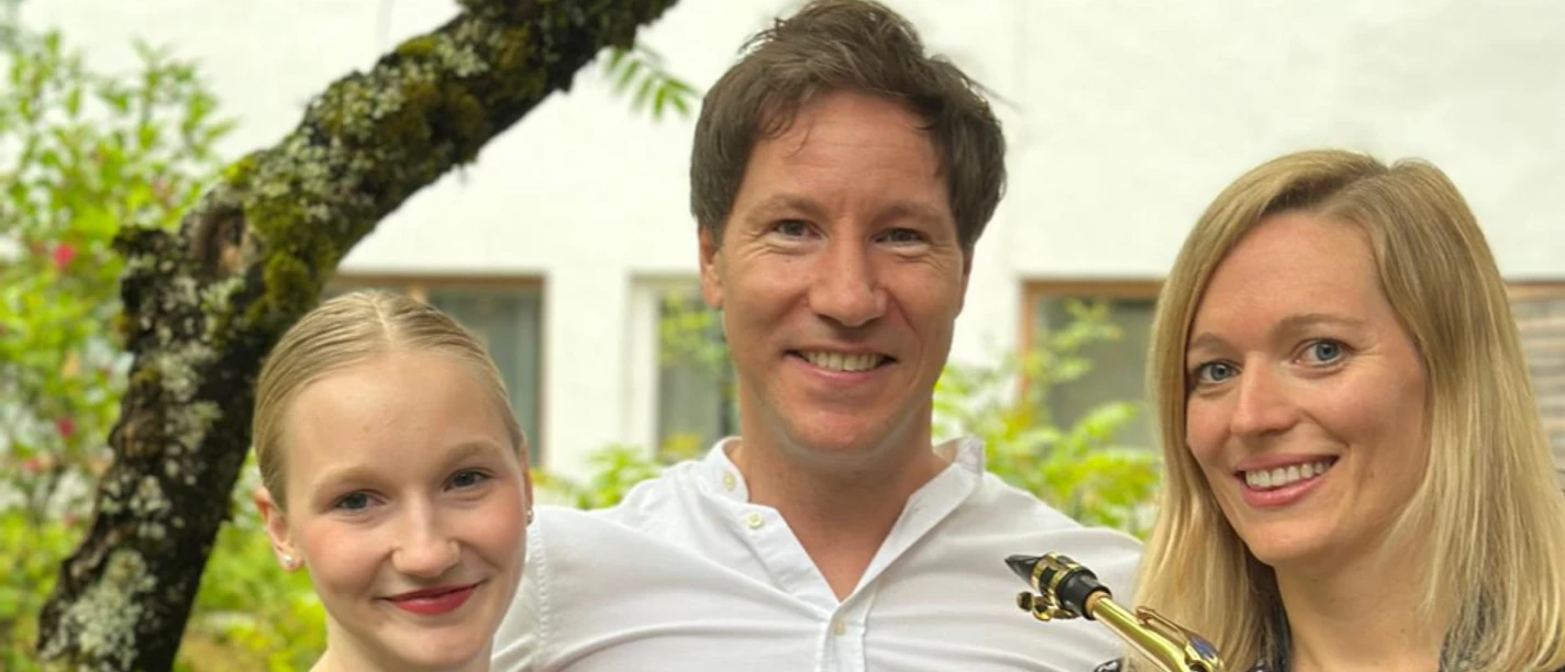 Summer Sounds.png Drei lachende Musiker, ein Mann und zwei Frauen, stehen draußen nebeneinander. Eine hält ein Saxophon.