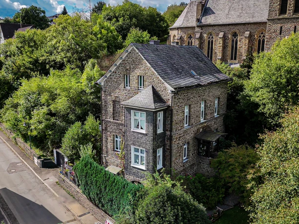 Katholisches Pfarrhaus Katholisches Pfarrhaus aus Stein mit Erker und Schieferdach, umgeben von Bäumen und einer Kirche.