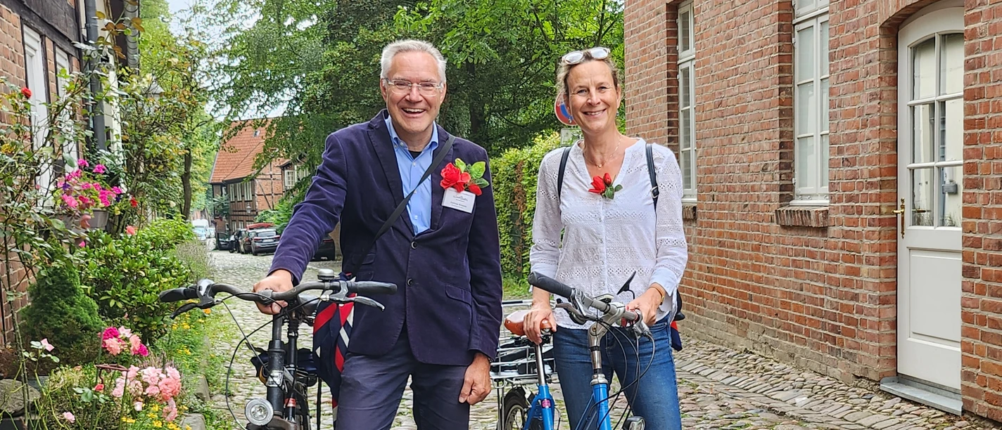 Rote Rosen-Radtour_CC BY-SA_©Verein Lüneburger Stadtführer.jpg Zwei Personen mit Fahrrädern und Rosen am Revers stehen auf einer Pflasterstraße. Sie Leiten eine Radtour zu Außendrehorten der Telenovela "Rote Rosen".