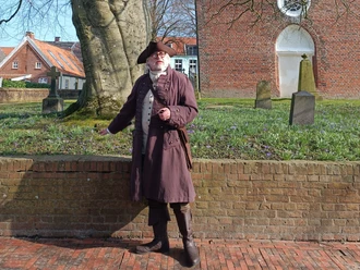 Hafenmeister Quirinus in Greetsiel Hafenmeister Quirinus im historischen Gewand mit Dreispitz vor der alten Backsteinkirche.