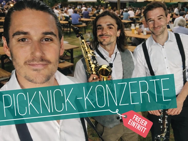 Picknick-Konzerte 2026 | 500 smile (Walking Act) Drei Musiker vor Bierbänken beim Open-Air-Picknickkonzert, Publikum im Hintergrund