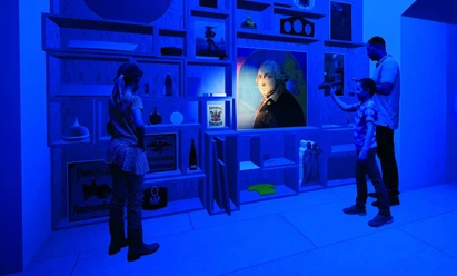 Auf dem Bild sieht man ein Regal voll mit Bildern, welches in dunkelblau gehalten ist und 2 Menschen mit einer Taschenlampe, welche auf ein Bild im Regal scheinen