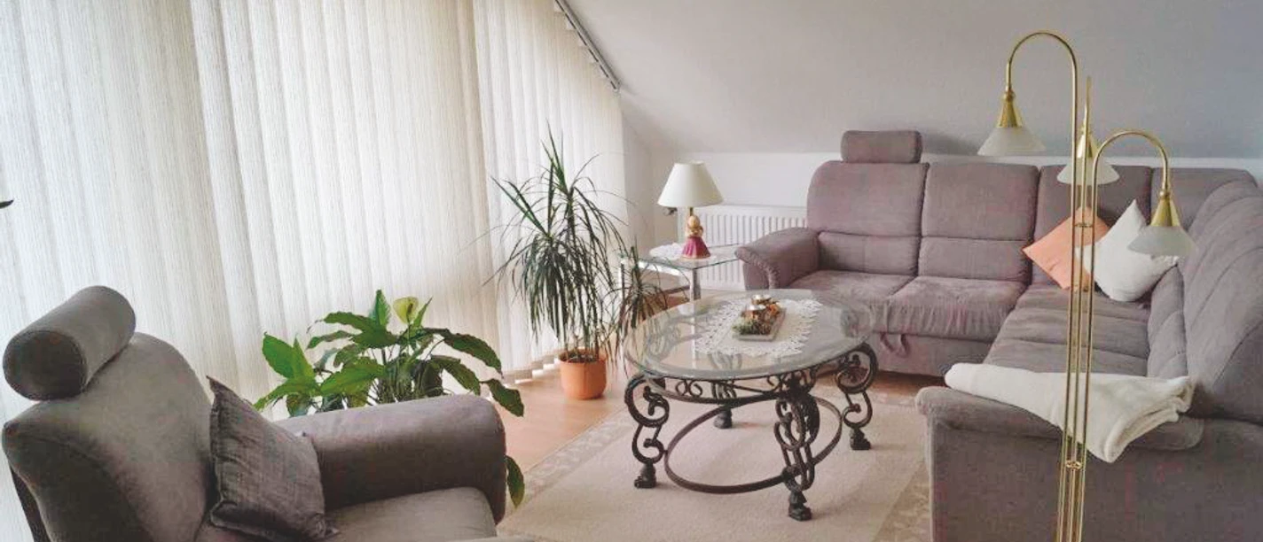 Ferienwohnung Pallme Wohnzimmer Helles Wohnzimmer mit grauem Ecksofa, Sessel, Glastisch und Zimmerpflanzen am großen Fenster.