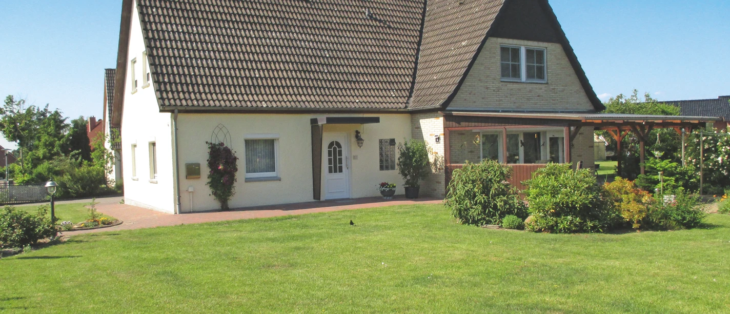 Ferienwohnung Schukat Außenansicht Helles Ferienhaus mit dunklem Dach, Terrasse und großer Rasenfläche unter blauem Himmel.