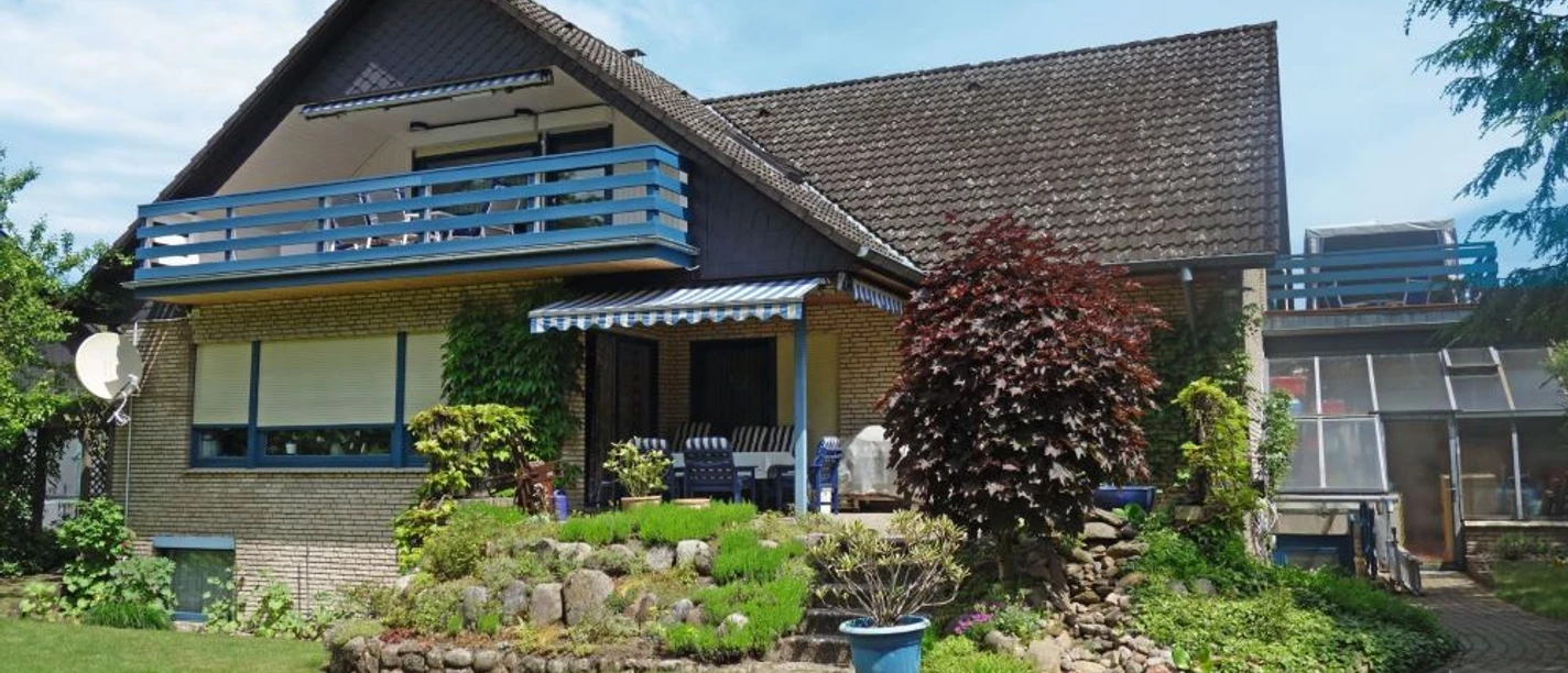 Haus Waldblick Außenansicht Klinkerhaus mit blauem Balkon, Terrasse und grünem Garten unter blauem Himmel.
