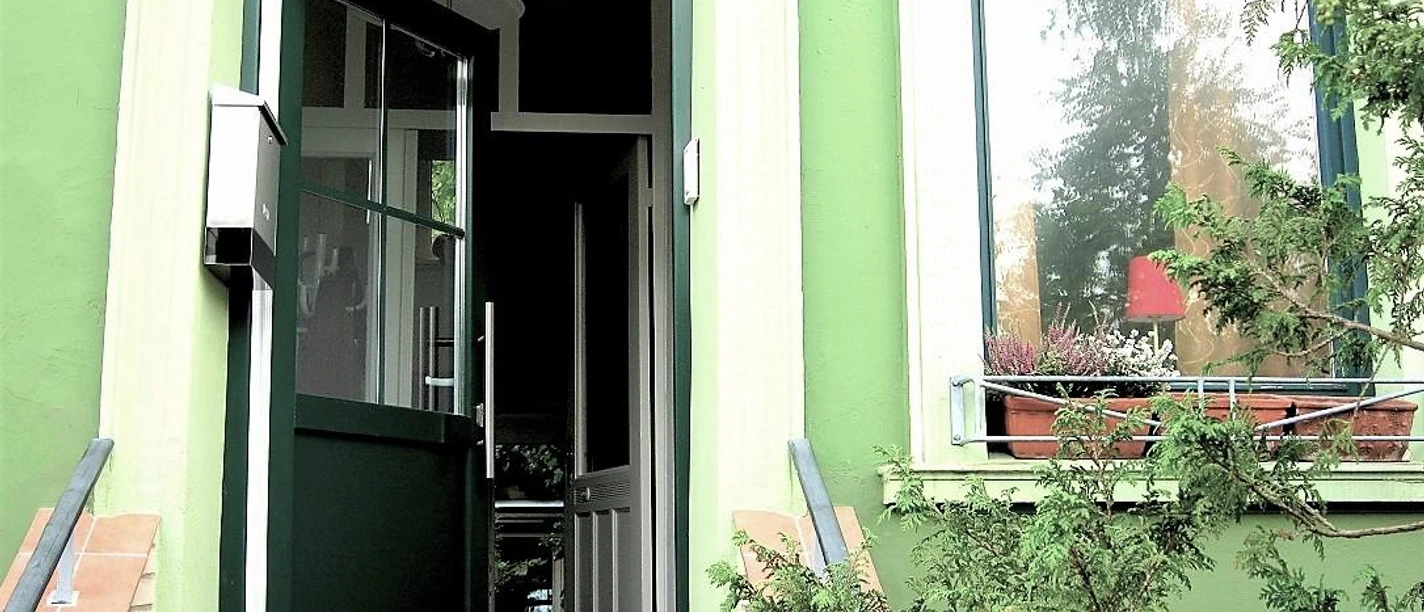 Kamps Ferienwohnungen Haupteingang Eingangstür eines hellgrünen Hauses mit der Hausnummer 58, daneben Fenster und Pflanzen.