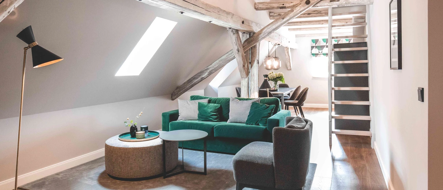 Saltbloom Apartments Wohnbereich Helles Dachgeschoss-Wohnzimmer mit freiliegenden Holzbalken, grünem Sofa und Essbereich.