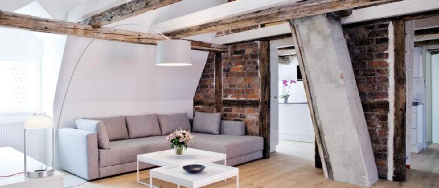 Schrangen Suites 1389 Wohnzimmer Helles Wohnzimmer mit historischem Fachwerk, Backsteinwand, grauem Ecksofa und weißen Tischen.