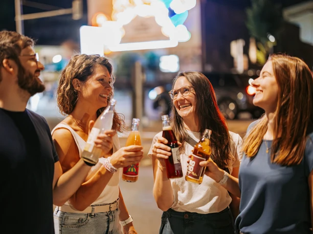 Nachtleben auf der KarLi - Szenemeilen in Leipzig Eine Gruppe Erwachsener steht abends mit Getränken in der Hand vor dem Leuchtschild "Löffelfamilie" an der Feinkost auf der Karl-Liebknecht-Straße