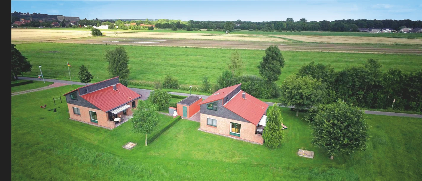 Wiesenhof Außenansicht Zwei rote Ziegeldach-Ferienhäuser auf einer weiten grünen Wiese mit Blick auf Felder und Bäume.