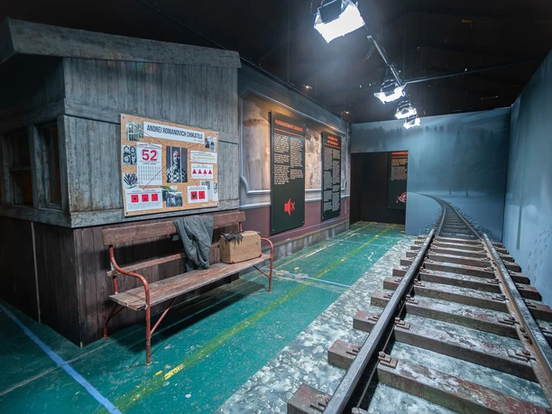 Serienkiller - Die True Crime Ausstellung Ausstellungsraum mit hölzerner Hütte, Bank und nachgebildetem Gleis in düsterer Szenerie.