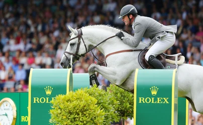 Ein weißes Pferd mit elegant gekleidetem Reiter springt über ein Hindernis mit dem Rolex-Logo. Im Hintergrund ist ein Publikum unscharf.