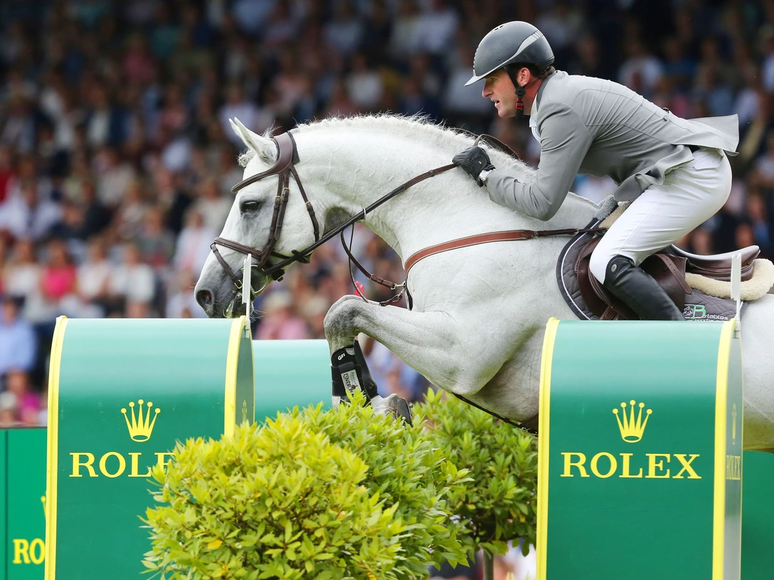 Ein weißes Pferd mit elegant gekleidetem Reiter springt über ein Hindernis mit dem Rolex-Logo. Im Hintergrund ist ein Publikum unscharf.
