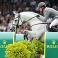 Ein weißes Pferd mit elegant gekleidetem Reiter springt über ein Hindernis mit dem Rolex-Logo. Im Hintergrund ist ein Publikum unscharf.