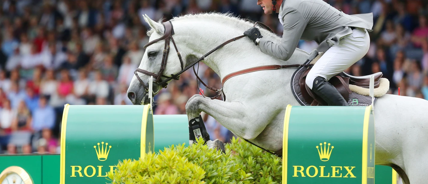 Ein weißes Pferd mit elegant gekleidetem Reiter springt über ein Hindernis mit dem Rolex-Logo. Im Hintergrund ist ein Publikum unscharf.