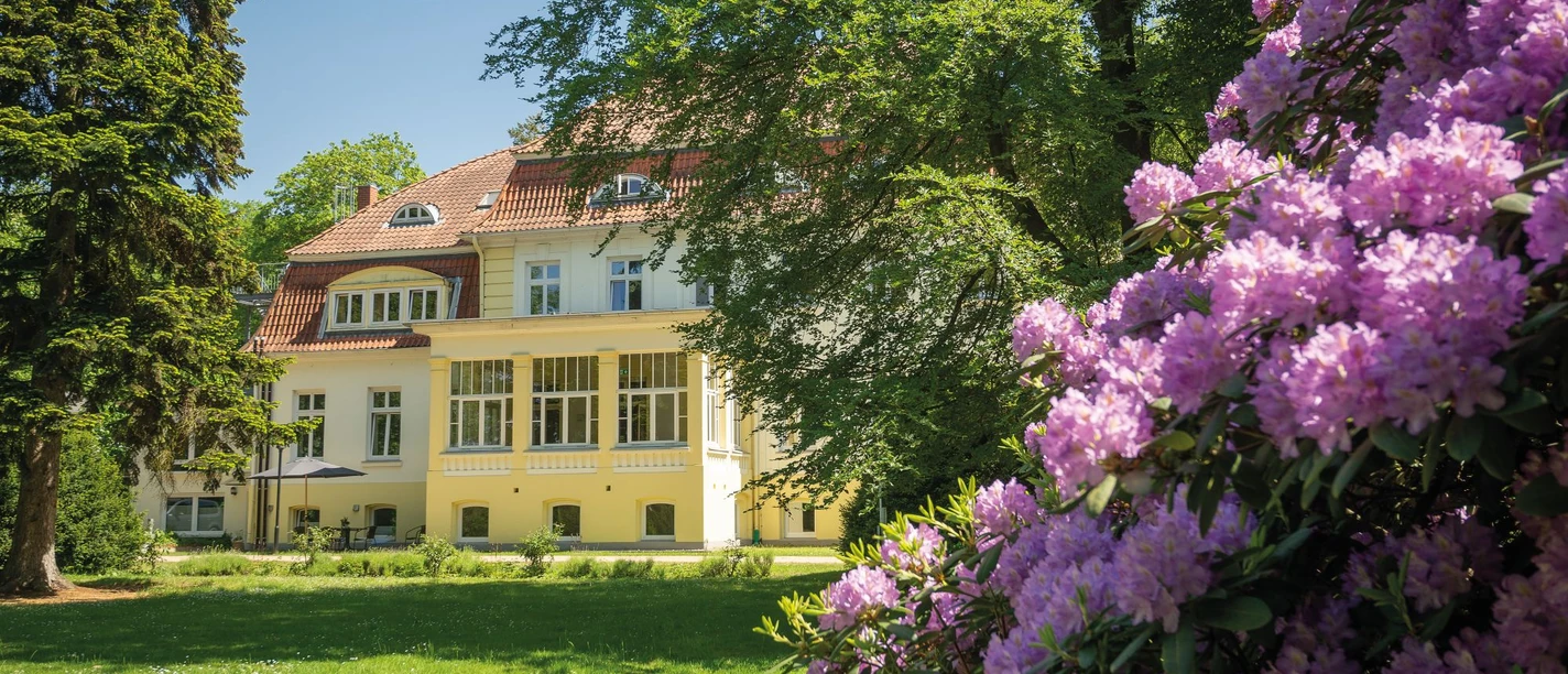 Gutshaus Barendorf Außenansicht Gutshaus Barendorf mit heller Fassade und rotem Dach, eingerahmt von lila Rhododendron.