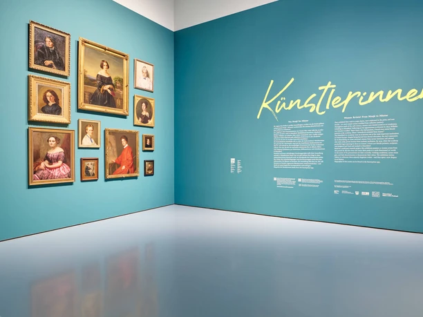 Künstlerinnen Porträtwand in Ausstellung mit gerahmten Bildern und großem Schriftzug Künstlerinnen.