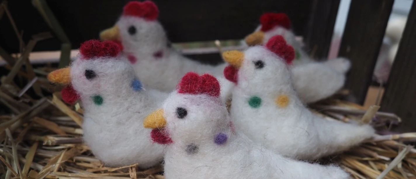 Gefilzte Hühner - MoorIZ Fünf handgemachte, weiße Filzhühner mit bunten Punkten und roten Kämmen sitzen auf trockenem Stroh.