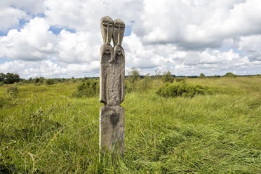 Ahlenmoor Totem Holzskulptur mit stilisierten Gesichtern steht in einer weiten, grünen Moorlandschaft unter Wolken.