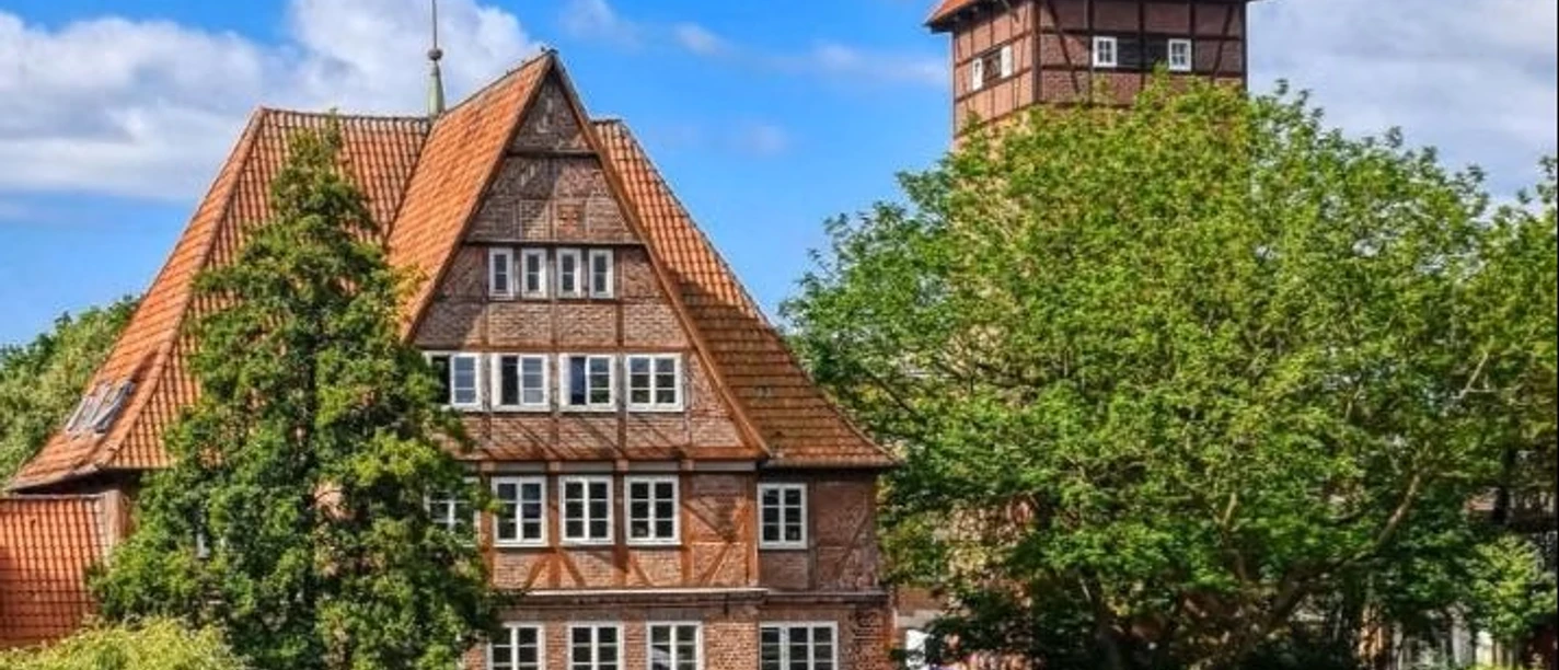 Hotel Ratsmühle Lüneburg Außenansicht Rotes Backsteingebäude der Ratsmühle mit Turm am Flussufer unter blauem Himmel in Lüneburg.