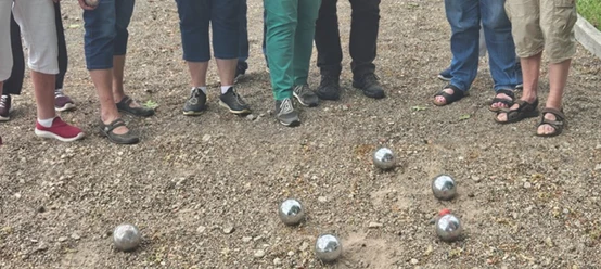 Eine Grupperuppe von Menschen, nur die Beine und Füße sind zu sehen, steht im Halbkreis um glänzende Boulekugeln auf einem Schotterplatz.