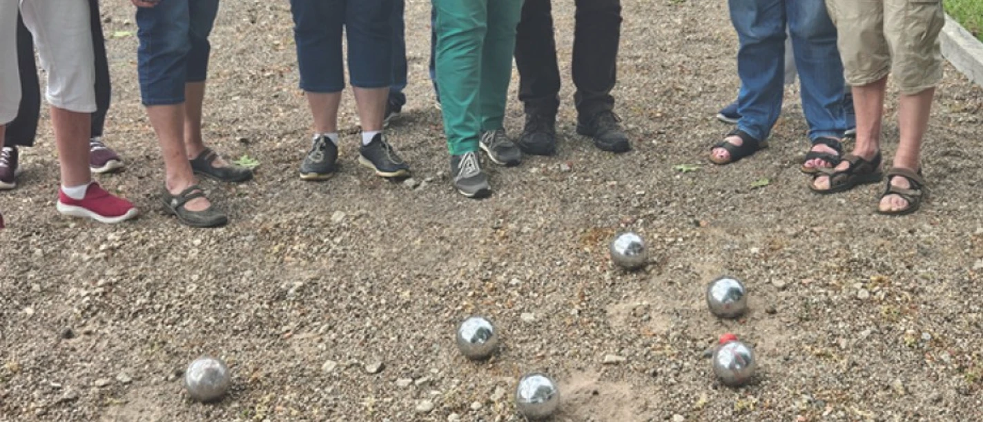 Eine Grupperuppe von Menschen, nur die Beine und Füße sind zu sehen, steht im Halbkreis um glänzende Boulekugeln auf einem Schotterplatz.