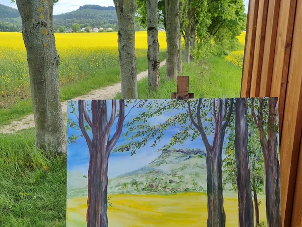 Draussen malen Sächsische-Schweiz mit Yvonne Brückner ein angefangenes Bild auf Staffelei im Vordergrund, Bäume, Weg und Rapsfeld. Im Hintergrund der Ort Gohrisch mit Tafelbergen.