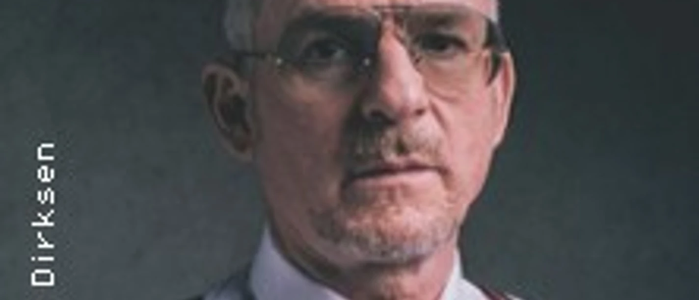 Älterer Mann mit weißem Haar, Brille, Hemd und Hosenträgern vor dunklem Hintergrund.