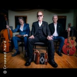 Drei Musiker sitzen mit Akkordeon, Cello und Gitarre in einem holzgetäfelten Raum.