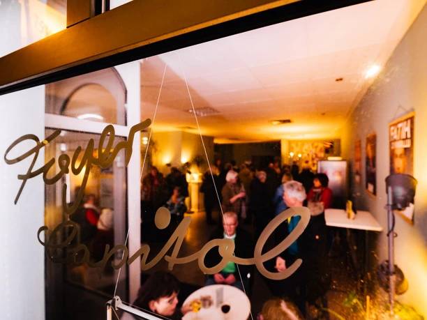 ANK_Capitol_BRHV_10MAR23-0205622 (c) Jonas Ginter Arbeitnehmerkammer Bremen.jpg Blick durch eine Glastür mit Aufschrift Foyer Capitol auf Menschen im beleuchteten Raum.