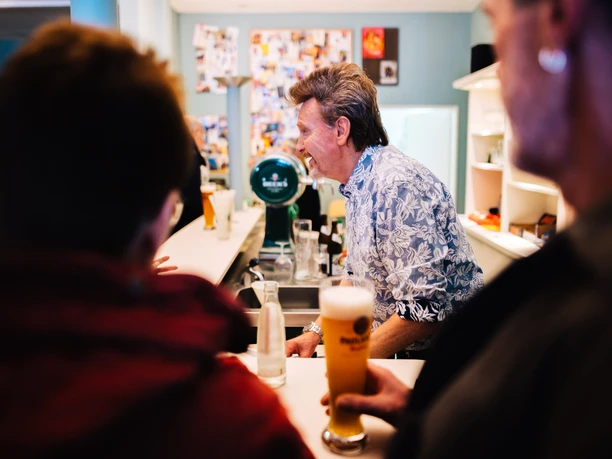 ANK_Capitol_BRHV_10MAR23-0405735 (c) Jonas Ginter Arbeitnehmerkammer Bremen.jpg Ein lächelnder Barkeeper im floralen Hemd bedient Gäste an der Theke im Capitol Bremerhaven.