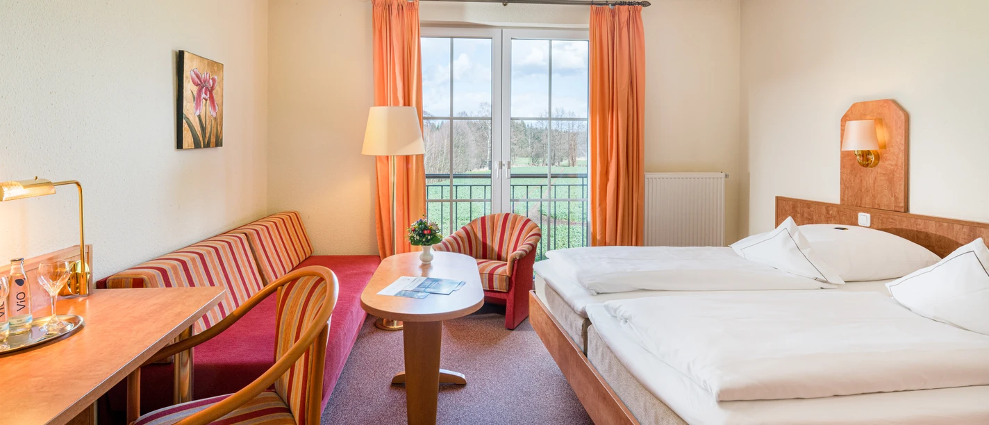 Wassermühle Heiligenthal Zimmeransicht Helles Hotelzimmer mit Doppelbett, rotem Sofa, Sitzecke und Blick ins Grüne.