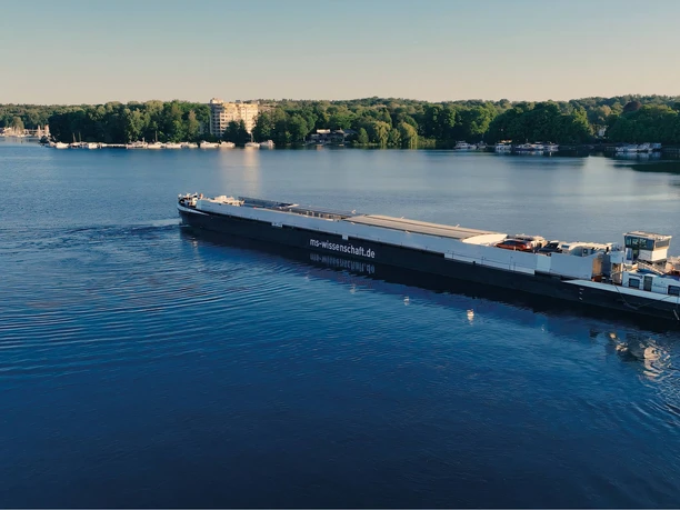MS Wissenschaft unterwegs auf Flüssen und Kanälen Ein langes Ausstellungsschiff gleitet über ruhiges Wasser vor einer grünen Uferkulisse.