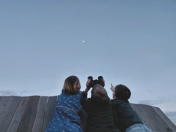 Gemeinde Hille Kinder Sternblick Drei Kinder stehen auf einem Holzsteg und beobachten mit Fernglas den Mond am Abendhimmel