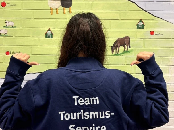 Lara.jpeg Rückenansicht einer jungen Frau im Pullover mit der Aufschrift "Team Tourismus-Service Butjadingen" vor einer Karte, die mit Symbolen die einzelnen Orte von Butjadingen zeigt.