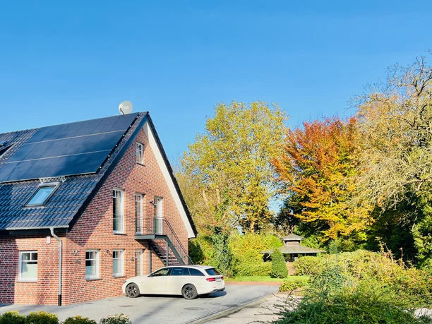 Apartment Hof Düker - Außenansicht Backsteinhaus mit Solardach, Außentreppe und Parkplatz vor Garten und Bäumen
