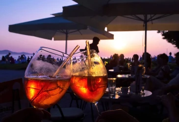 Zwei Aperol-Gläser Zwei Aperolgläser stoßen im Abendlicht an, umgeben von Sitzplätzen und sanfter Silhouette.