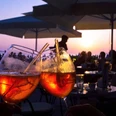 Zwei Aperol-Gläser Zwei Aperolgläser stoßen im Abendlicht an, umgeben von Sitzplätzen und sanfter Silhouette.