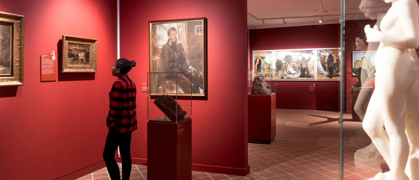 Gemäldeabteilung_Ostpreussisches_Landesmuseum.jpeg Person betrachtet Gemälde im Ostpreußischen Landesmuseum in einem Raum mit roten Wänden. Im Vordergrund steht eine Statue.