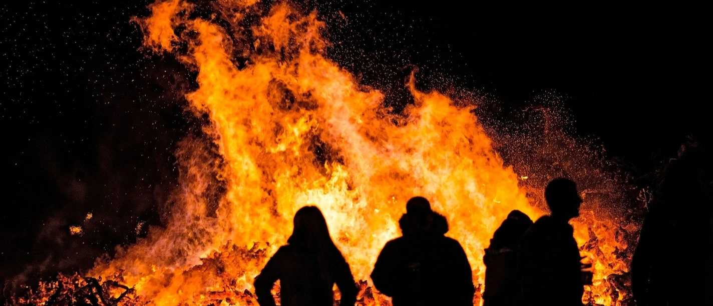 Osterfeuer-Burlage.jpg Silhouetten von Menschen betrachten nachts ein großes, hell loderndes Osterfeuer.