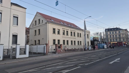 Vue latérale de la façade de la maison Erich Zeigner à Leipzig au crépuscule, avec la rue et les rails du tramway devant elle.