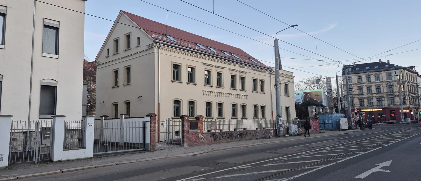 Vue latérale de la façade de la maison Erich Zeigner à Leipzig au crépuscule, avec la rue et les rails du tramway devant elle.