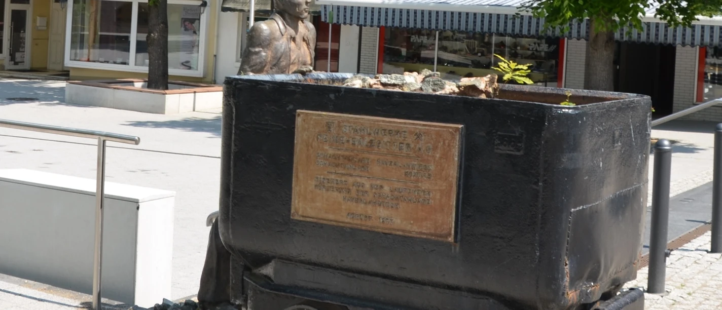Bergmann_HD_16zu9.JPG Bronzestatue eines Bergmanns hinter einer schwarzen Lore mit Gedenktafel am Marktplatz.