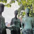 Drei Grazien_HD_16zu9.jpg Drei stehende Frauenstatuen aus Bronze mit grüner Patina unter den Blättern eines Baumes.