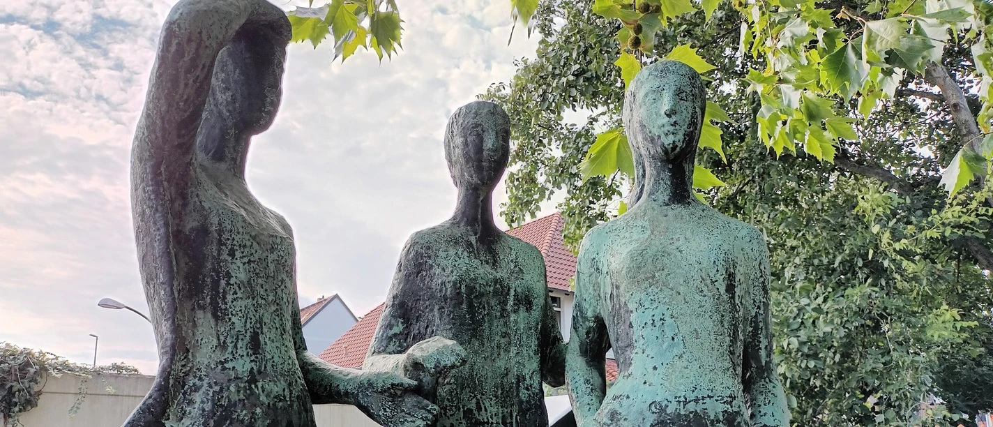 Drei Grazien_HD_16zu9.jpg Drei stehende Frauenstatuen aus Bronze mit grüner Patina unter den Blättern eines Baumes.