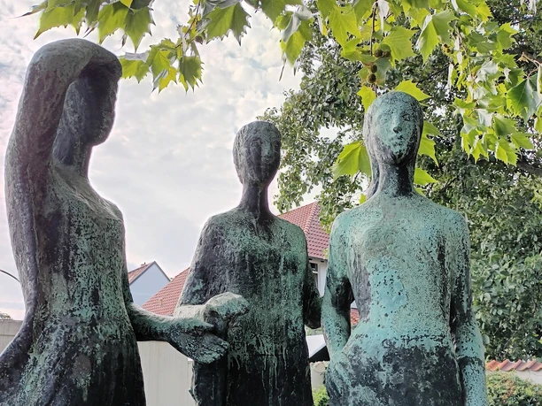Drei Grazien_HD_16zu9.jpg Drei stehende Frauenstatuen aus Bronze mit grüner Patina unter den Blättern eines Baumes.