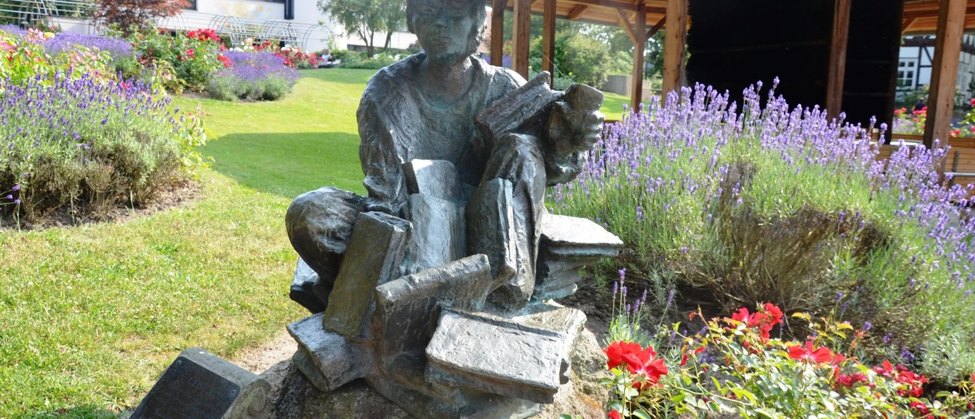 Lesende_HD_16zu9.jpg Bronzestatue Die Lesende auf Büchern in einem Garten mit Lavendel und roten Rosen.